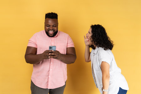 Young couple in casual clothing standing together, woman desperately screaming near happy man using phone and ignoring her, attracting his attention. Indoor studio shot isolated on yellow background.の写真素材