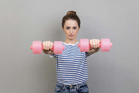 Portrait of serious concentrated woman wearing striped T-shirt holding out pink dumbbells to camera, working out alone. Indoor studio shot isolated on gray background.の写真素材
