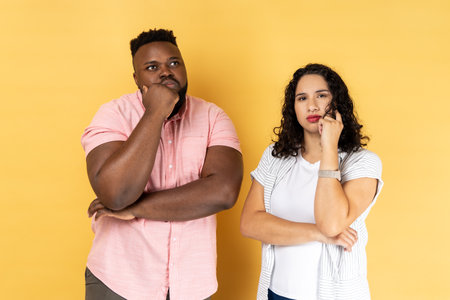 Portrait of thoughtful young couple in casual clothing standing together, holding their chins while thinking solution, looking away. Indoor studio shot isolated on yellow background.の写真素材