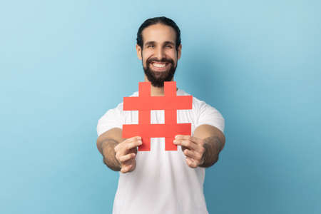 Portrait of smiling happy man with beard wearing white T-shirt holding out hashtag symbol, recommending popular topics, internet trends. Indoor studio shot isolated on blue background.の写真素材