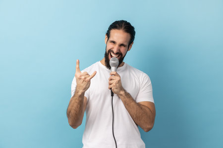 Portrait of excited man with beard in white T-shirt loudly singing song holding microphone in hand, having fun resting in karaoke, showing rock sign. Indoor studio shot isolated on blue background.の写真素材