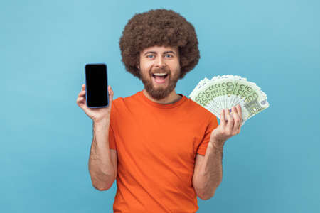 Portrait of satisfied man with Afro hairstyle wearing orange T-shirt showing euro banknotes and smart phone with empty display for promotional text. Indoor studio shot isolated on blue background.の写真素材