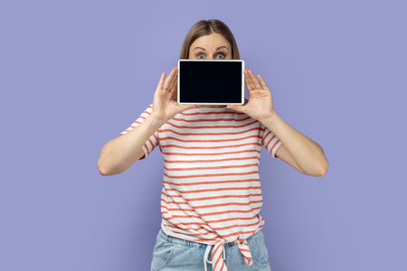 Portrait of shocked astonished blond woman wearing striped T-shirt covering half of face with...
