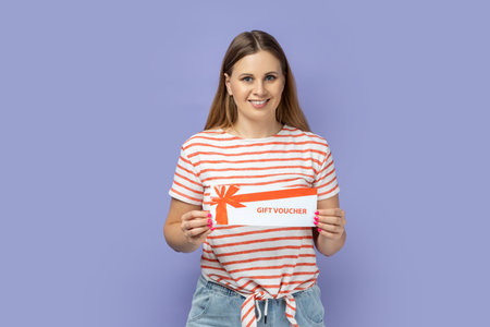 Portrait of smiling happy pretty blond woman wearing striped T-shirt holding voucher for gift,...