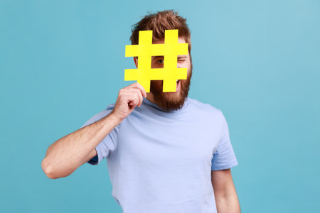 Portrait of bearded man hiding face behind yellow hashtag sign, calls on to tag topics on websites,...