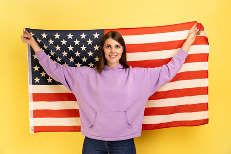 Portrait of attractive happy positive woman standing with raised arms, holding USA flag, celebrating national holiday, wearing purple hoodie. Indoor studio shot isolated on yellow background.の写真素材