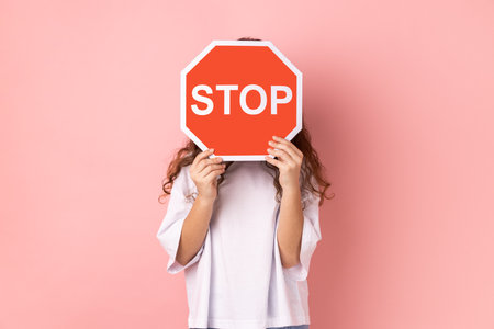 Portrait of anonymous little girl wearing white T-shirt covering face with stop symbol, holding red traffic sign, forbidden access. Indoor studio shot isolated on pink background.の写真素材