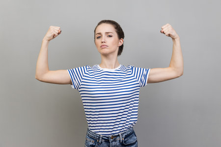 Portrait of proud woman wearing striped T-shirt showing biceps, expressing power ambitions to become successful, female rights, feminism. Indoor studio shot isolated on gray background.の写真素材