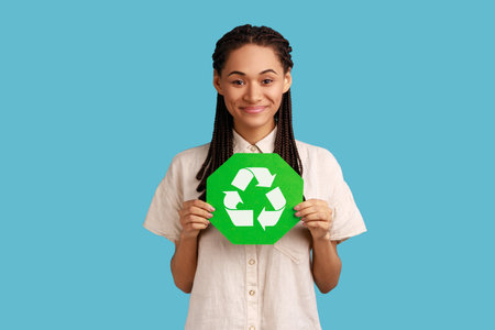 Responsible self confident woman with black dreadlocks holding green recycling sign in hand and seriously looking at camera, thinking green. Indoor studio shot isolated on blue background.の写真素材