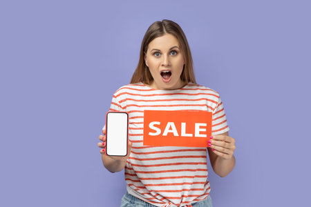 Portrait of astonished surprised blond woman wearing striped T-shirt holding sale card and smart phone with empty display, copy space for promotion. Indoor studio shot isolated on purple background.の写真素材