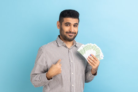Portrait of young adult businessman with beard holding euro bills and pointing at himself with...