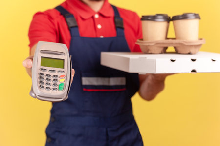 Anonymous delivery man standing with pizza box and coffee in disposable cup and showing pos terminal for online paying for delivery. Indoor studio shot isolated on yellow backgroundの写真素材