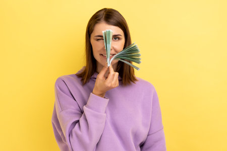 Portrait of young adult dark haired woman standing looking throw euro banknotes, looking at camera with one eye, wearing purple hoodie. Indoor studio shot isolated on yellow background.の写真素材