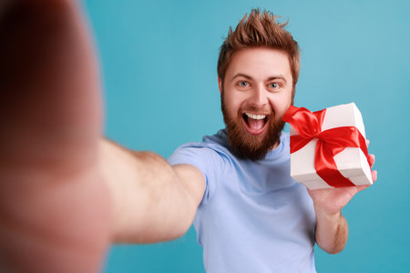 Portrait of positive bearded man with widely opened mouth looking at camera with happy smile POV, point of view of photo, holding present box. Indoor studio shot isolated on blue background.の写真素材