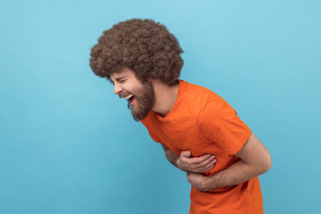 Side view of laughing man with Afro hairstyle in orange T-shirt holding his stomach and hunched in crazy hysterical laughter, sincere joyful emotions. Indoor studio shot isolated on blue background.の写真素材