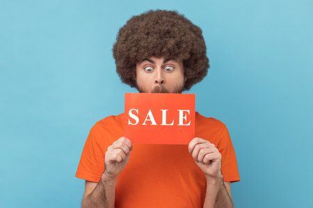 Man with Afro hairstyle wearing orange T-shirt standing with Sale word, looking with big eyes surprised, shocked by discounts, Black Friday low prices. Indoor studio shot isolated on blue background.の写真素材