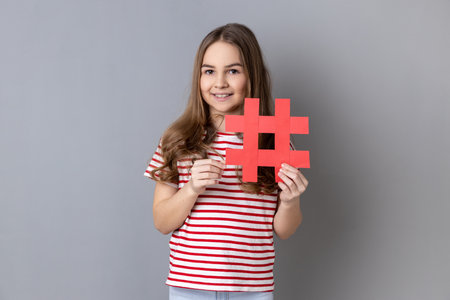 Portrait of delighted positive dark haired little girl wearing striped T-shirt holding red big paper hash symbol and smiling at camera. Indoor studio shot isolated on gray background.の写真素材