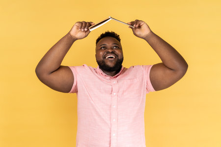 Portrait of joyful delighted bearded man wearing pink shirt holding opened book on head, looking up with happy positive expression. Indoor studio shot isolated on yellow background.の写真素材