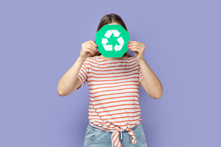 Portrait of unknown anonymous woman wearing striped T-shirt holding green recycling sign and hiding her face behind ecology symbol. Indoor studio shot isolated on purple background.の写真素材