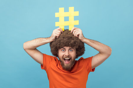 Portrait of man with Afro hairstyle holding yellow hashtag sign board on head, dreaming about target tagging posts on his website, smm manager. Indoor studio shot isolated on blue background.の写真素材