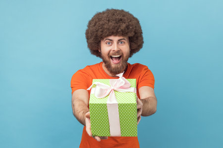 Portrait of man with Afro hairstyle wearing orange T-shirt giving gift, congratulating on birthday and offering surprise, holiday bonus wrapped in box. Indoor studio shot isolated on blue background.の写真素材
