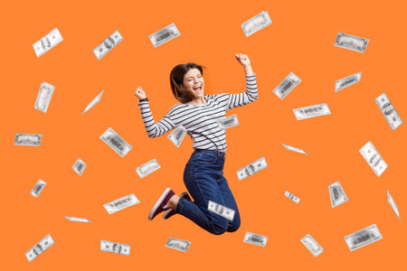 Portrait of satisfied excited woman jumping with clenched fist, celebrating winning lottery, flying in money rain, dollars falling. Indoor studio shot isolated on orange background.の写真素材