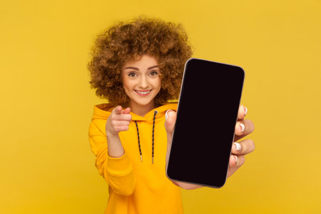 Portrait of attractive smiling woman with Afro hairstyle wearing hoodie showing big mobile display for copy space and pointing to camera, choosing you. Indoor studio shot isolated on yellow backgroundの写真素材
