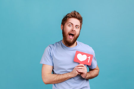 Portrait of excited bearded man holding social media heart Like button in front his chest, emoji counter, follower notification. Indoor studio shot isolated on blue background.の写真素材