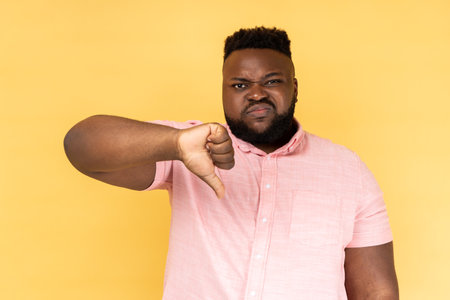Portrait of sad upset bearded man wearing pink shirt showing thumb down, expressing bad negative emotions, dislike, frowning face. Indoor studio shot isolated on yellow background.の写真素材