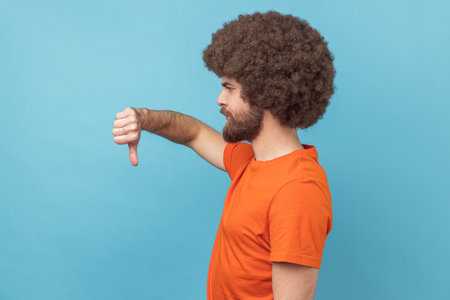 Side view of displeased man with Afro hairstyle wearing orange T-shirt showing thumbs down dislike...