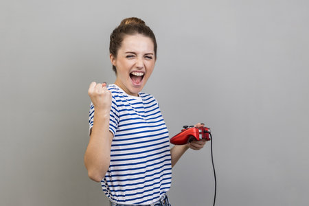 Portrait of woman wearing striped T-shirt holding gamepad, winning video games competition, clenched fist with happy expression, rejoices victory. Indoor studio shot isolated on gray background.の写真素材