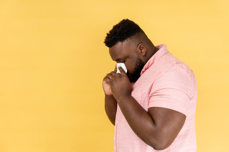 Side view portrait of man wearing pink shirt hiding face in hand with white handkerchief, crying, feeling stressed, worried facial expression. Indoor studio shot isolated on yellow background.の写真素材