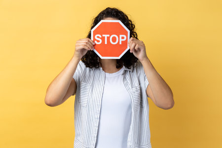 Anonymous woman with dark wavy hair standing covering her face with red stop sign, warning about...