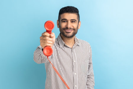 Take it, answer call. Man giving phone handset looking at camera, smiling looking friendly joyful, helpful contact center, wearing striped shirt. Indoor studio shot isolated on blue background.の写真素材