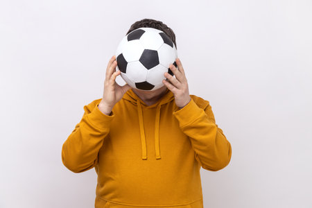 Anonymous unknown man hiding behind soccer ball, football fan covering his face during match, wearing urban style hoodie. Indoor studio shot isolated on white background.の写真素材