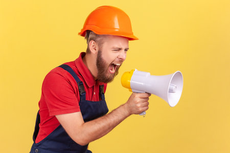 Side view of angry bearded worker wearing protective helmet and blue overalls holding megaphone and screaming with aggressive expression, protesting. Indoor studio shot isolated on yellow background.の写真素材