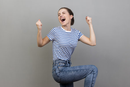 I am champion. Portrait of woman wearing striped T-shirt screaming for joy, raising high her fists, celebrating successful winning, incredible victory. Indoor studio shot isolated on gray background.の写真素材