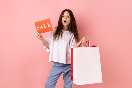 Portrait of shocked little girl wearing white T-shirt holding shopping bags and sale card, astonished with big holidays discounts. Indoor studio shot isolated on pink background.の写真素材