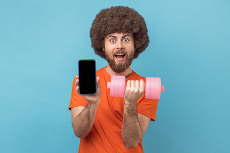 Portrait of shocked man with Afro hairstyle wearing orange T-shirt holding dumbbell and smart phone with empty display for advertisement. Indoor studio shot isolated on blue background.の写真素材