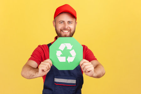 Portrait of smiling happy worker man standing and holding recycling sign, thinking green, looking at camera, wearing overalls and red cap. Indoor studio shot isolated on yellow background.の写真素材