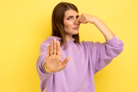 Portrait of woman grimacing with disgust, holding breath, pinching nose with fingers to avoid bad smell, showing stop gesture, wearing purple hoodie. Indoor studio shot isolated on yellow backgroundの写真素材