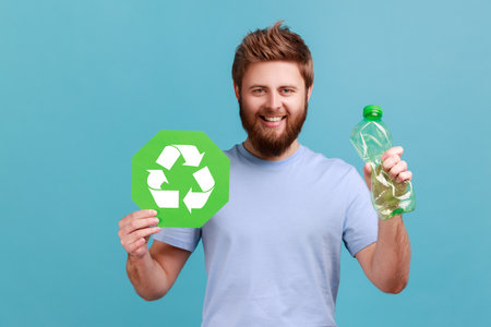 Portrait of smiling optimistic handsome bearded man pressing bottle holding in hand recycling sign board, plastic disposal, garbage sorting. Indoor studio shot isolated on blue background.の写真素材