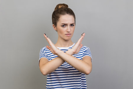 No way, absolutely not. Portrait of woman wearing striped T-shirt showing x sign with crossed hands, meaning stop, this is the end. Indoor studio shot isolated on gray background.の写真素材