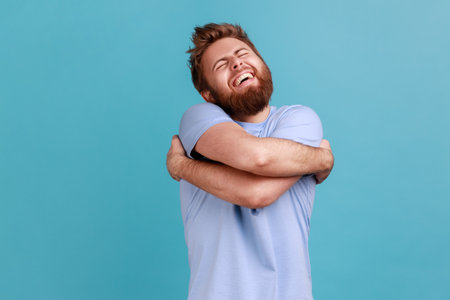 Portrait of satisfied positive handsome bearded man embracing himself, closed eyes and smiling with pleasure, complacency and egoism. Indoor studio shot isolated on blue background.の写真素材
