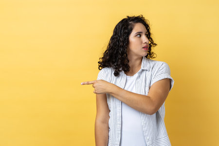 Portrait of woman with dark wavy hair pointing finger aside, ordering get out and looking resentful, boss dismissing from work, showing exit. Indoor studio shot isolated on yellow background.の写真素材