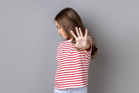 Portrait of scared serious little dark haired girl wearing striped T-shirt showing block stop gesture, set back bullying and violence. Indoor studio shot isolated on gray background.の写真素材