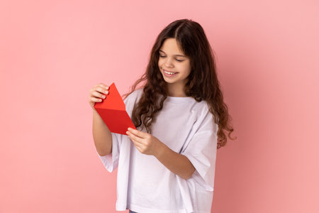 Portrait of little girl wearing white T-shirt reading letter or greeting card, holding envelope, smiling and rejoicing pleasant news. Indoor studio shot isolated on pink background.の写真素材