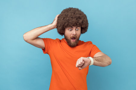Portrait of man with Afro hairstyle wearing orange T-shirt pointing finger on his wrist watch with nervous face, time is out, keeps hand on head. Indoor studio shot isolated on blue background.の写真素材