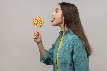 Side view of childish woman licking multicolor candy, wants to eat, looking at camera, showing...