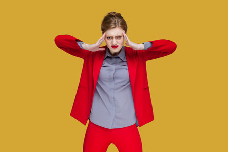 Portrait of sad upset unhealthy woman with red lips standing with hands on temples, suffering terrible pain, having headache, wearing red jacket. Indoor studio shot isolated on yellow background.の写真素材